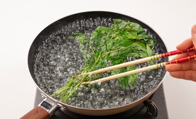 輪ゴムで束ねたモロヘイヤをフライパンに入れ、箸で押さえながらゆでている写真