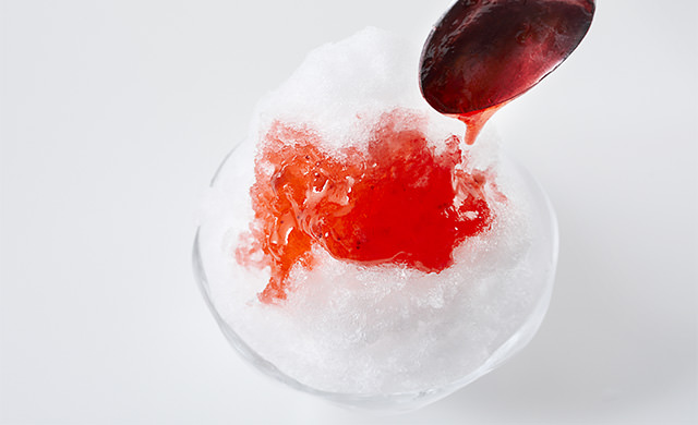 三角形に盛りつけたかき氷にスプーンでいちごシロップをかけている写真